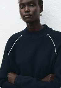 Marineblauer Strickpullover mit hohem Kragen und geripptem Muster, mit weißen Akzentnähten entlang der Schultern. Knöpfe nicht sichtbar.