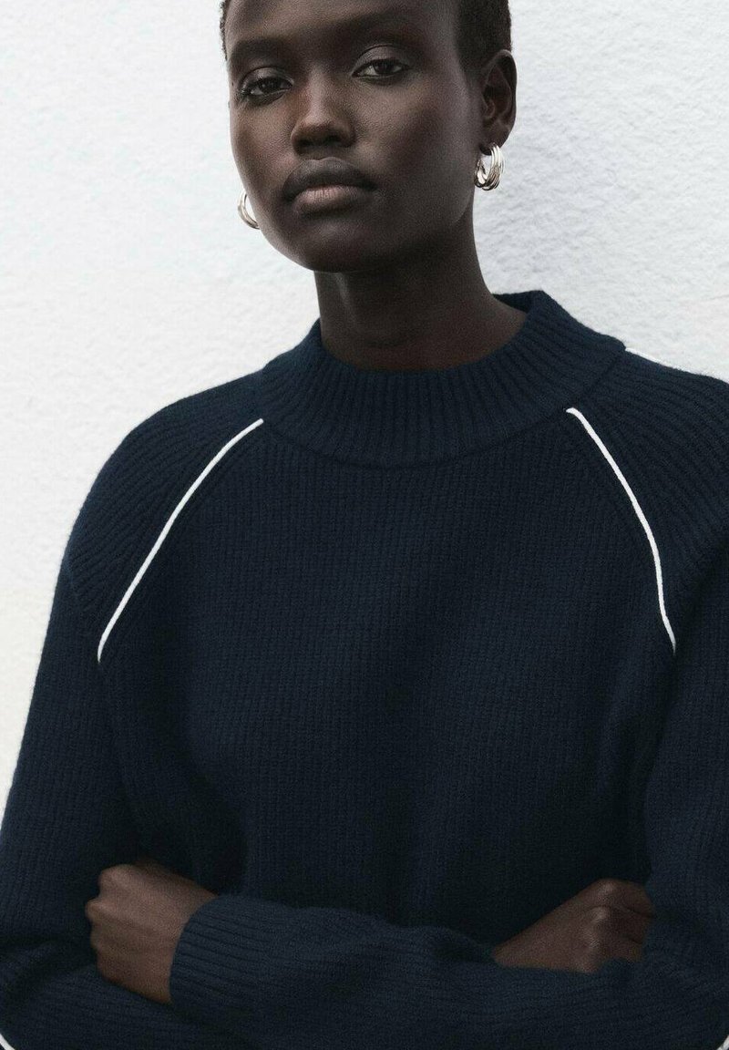 Marineblauer Strickpullover mit hohem Kragen und geripptem Muster, mit weißen Akzentnähten entlang der Schultern. Knöpfe nicht sichtbar.