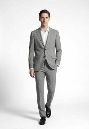 Mann geht nach vorne, trägt einen hellgrauen Zweiknopf-Anzug, weißes Hemd und schwarze Anzugsschuhe vor weißem einfarbigem Hintergrund.