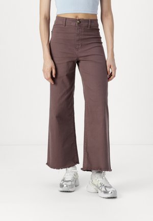 Weite, hoch taillierte Hosen aus hellbraunem Denim mit ausgefranstem Saum. Kombiniert mit weißen Sneakers mit grauen und silbernen Akzenten.