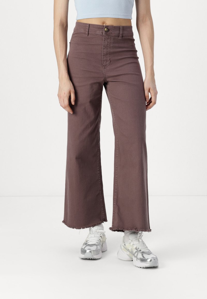 Pantalons larges taille haute en denim marron clair avec un ourlet effiloché. Associés à des baskets blanches avec des accents gris et argent.
