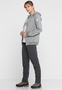 Sudadera con cremallera de color gris claro con un bolsillo tipo canguro y un logo en la manga, combinada con pantalones gris oscuro y zapatos de senderismo marrones con suelas negras.