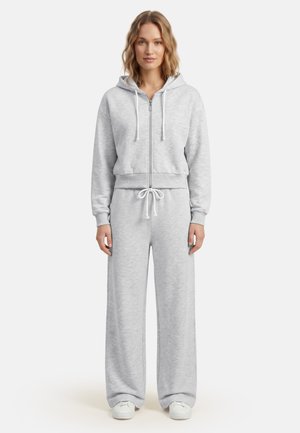 Vrouw die een lichtgrijze zip-up hoodie en bijpassende losse joggingbroek draagt met witte sneakers, staand tegen een witte achtergrond.
