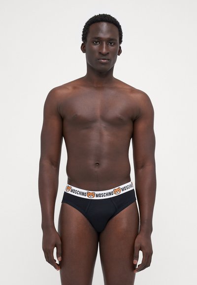 Fekete alsónadrág fehér elasztikus derékkal, amelyen egy medve grafika és a "MOSCHINO" szöveg található. Simább anyag és szoros illeszkedés.