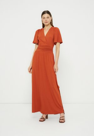 Woman wearing a burnt orange, short-sleeve, V-neck, floor-length dress with a fitted waist and brown strappy sandals, standing against a white background.