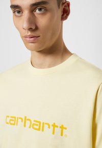 T-shirt de algodão amarelo com um grande logótipo laranja "carhartt" numa fonte moderna, com um clássico decote redondo e mangas curtas.