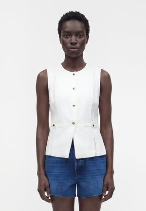 Femme portant un gilet blanc sans manches boutonné avec des accents dorés et un short en jean bleu, debout sur un fond blanc uni.