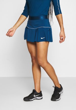 Osoba oblečená v modré sportovní sukni, odpovídajícím topu a černých teniskách Nike stojící před bílým pozadím.