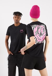Svarte T-skjorter har en liten rosa smiley på fronten og et stort rosa smiley-graphic med zebra-striper på baksiden. Modellen har på seg svarte shorts.