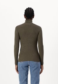 Personne avec des cheveux tressés portant un pull à col roulé côtelé vert olive et un jean bleu, se tenant de dos devant un fond blanc.