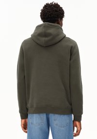 Felpa verde scuro in misto cotone, con tasca a marsupio frontale, cappuccio con cordino e polsini a costine. Indossata con jeans azzurri chiari.