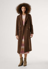 Brown wool blend double-breasted coat with large lapels, side pockets, and silver buttons, layered over a patterned dress and ankle boots.