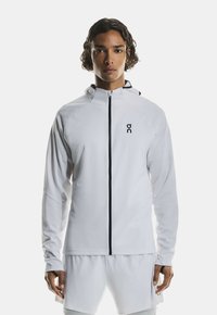 White athletic jacket with a high collar, black zipper, and logo on the chest. Smooth texture and long sleeves. Minimalist design.