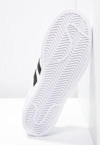 Bílá teniska s texturovanou gumovou podrážkou, černými detaily a klasickým designem se třemi pruhy. Boční pohled na světle pozadí.