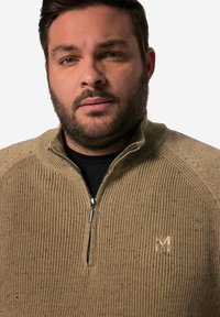 Beige zip-up tröja med ribbad textur, med spräckliga mönster och kontrasterande axelaccenter. Diskret logotyp på bröstet.