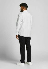 Weißes Langarmhemd mit Kragen, schwarze Hose und weiße Sneaker. Die Rückansicht zeigt Details an den Ärmeln und eine lockere Passform.