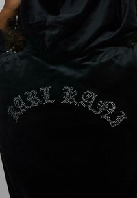 Crna baršunasta hoodie s kamenčićima koji oblikuju natpis "KARL KAP" smješten na leđima u zakrivljenom dizajnu. Glatka tekstura.