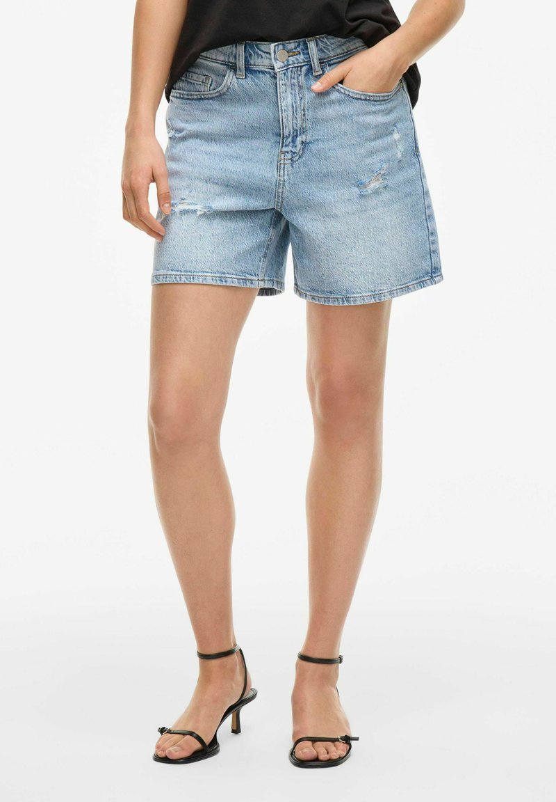 Shorts in denim blu chiaro con vita alta, taglio dritto e dettagli strappati. Abbinati a sandali neri con cinturini.