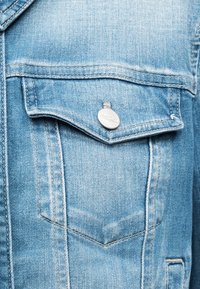 Τσέπη από γαλάζιο τζιν μπουφάν με καπάκι που ασφαλίζει με στρογγυλό ασημένιο κουμπί σε κοντινή άποψη.