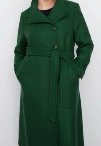 Cappotto verde con cintura, colletto, chiusura a un bottone, due tasche laterali e tessuto testurizzato. Vestibilità sartoriale con dettaglio della cintura in vita.