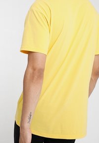 Camiseta de algodón amarilla de manga corta. Corte recto con una textura suave. Muestra la parte posterior y un tatuaje en la muñeca izquierda.
