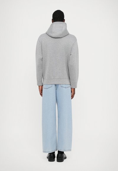 Grijs pullover hoodie met een ruime achterkant, gecombineerd met lichtblauwe wijdbenige jeans. Zwarte schoenen. Effen achtergrond.