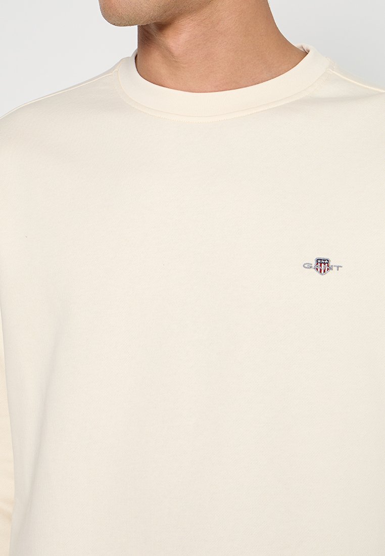 Beiger Sweatshirt mit Rundhalsausschnitt, weicher Textur und kleinem gesticktem Logo auf der Brust. Klassisches und vielseitiges Design.