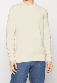 Hellbeige, gerippter Pullover mit Rundhalsausschnitt und langen Ärmeln, versehen mit einem strukturierten Strickmuster, kombiniert mit blauen Jeans.