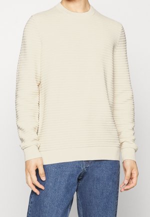 Hellbeige, gerippter Pullover mit Rundhalsausschnitt und langen Ärmeln, versehen mit einem strukturierten Strickmuster, kombiniert mit blauen Jeans.