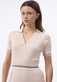 Gebreide polokleding in lichtbeige met een kabelstructuur, korte mouwen, een V-hals en contrasterende gestreepte accenten bij de taille en mouwmanchetten.