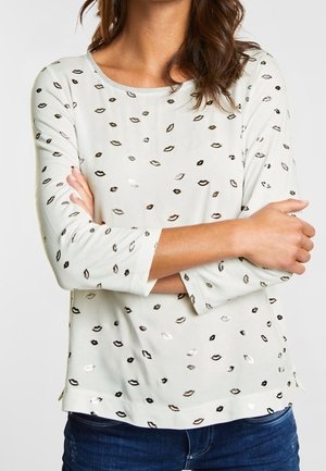 Mujer con una camisa blanca de manga larga con estampados de labios en negro, marrón y plata, brazos cruzados, combinada con vaqueros azules.