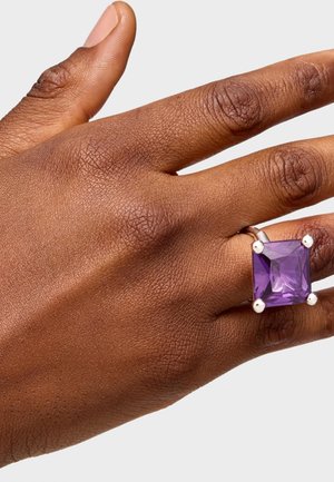 Anillo de gema morada cortado en cuadrado, engastado en plata, que presenta cuatro garras y un acabado pulido, mostrado en una mano con piel texturizada.