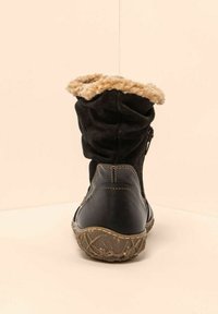 Botín de invierno negro con forro de lana de color beige, cremallera lateral y suela texturizada, mostrado desde atrás sobre un fondo beige.