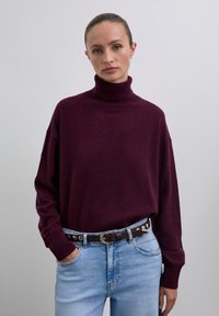 Woman wearing a dark purple turtleneck sweater tucked into light blue jeans with a dark brown studded belt, standing against a plain background.