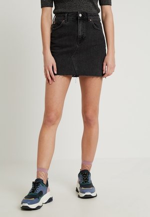 Personen har en sort denim mininederdel med rå kant, grå top, pink netstrømper og chunky multifarvede sneakers på og står på en lys baggrund.
