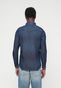 Tumši zila džinsa krekls ar garām piedurknēm, pogai uz aprocēm, asim apkakli un izliektu apakšu. Raksturīga muguras jakas daļa un divas aizmugurējās kabatas.