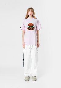 Camiseta oversized de color rosa claro con un diseño de oso de peluche marrón texturizado, combinada con pantalones blancos y zapatos grises sin cordones.