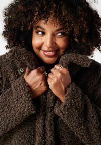 Bruine textuur jas met een fuzzy, krullend stof. Heeft een knoopdetail op de mouw. Model houdt de jas dicht bij de kraag.