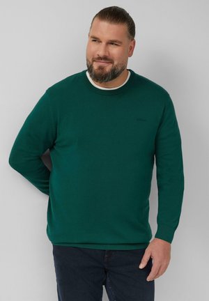 Maglione verde con collo rotondo, polsini e orlo a costine. Realizzato in tessuto testurizzato. Indossato sopra una camicia bianca, abbinato a jeans scuri.