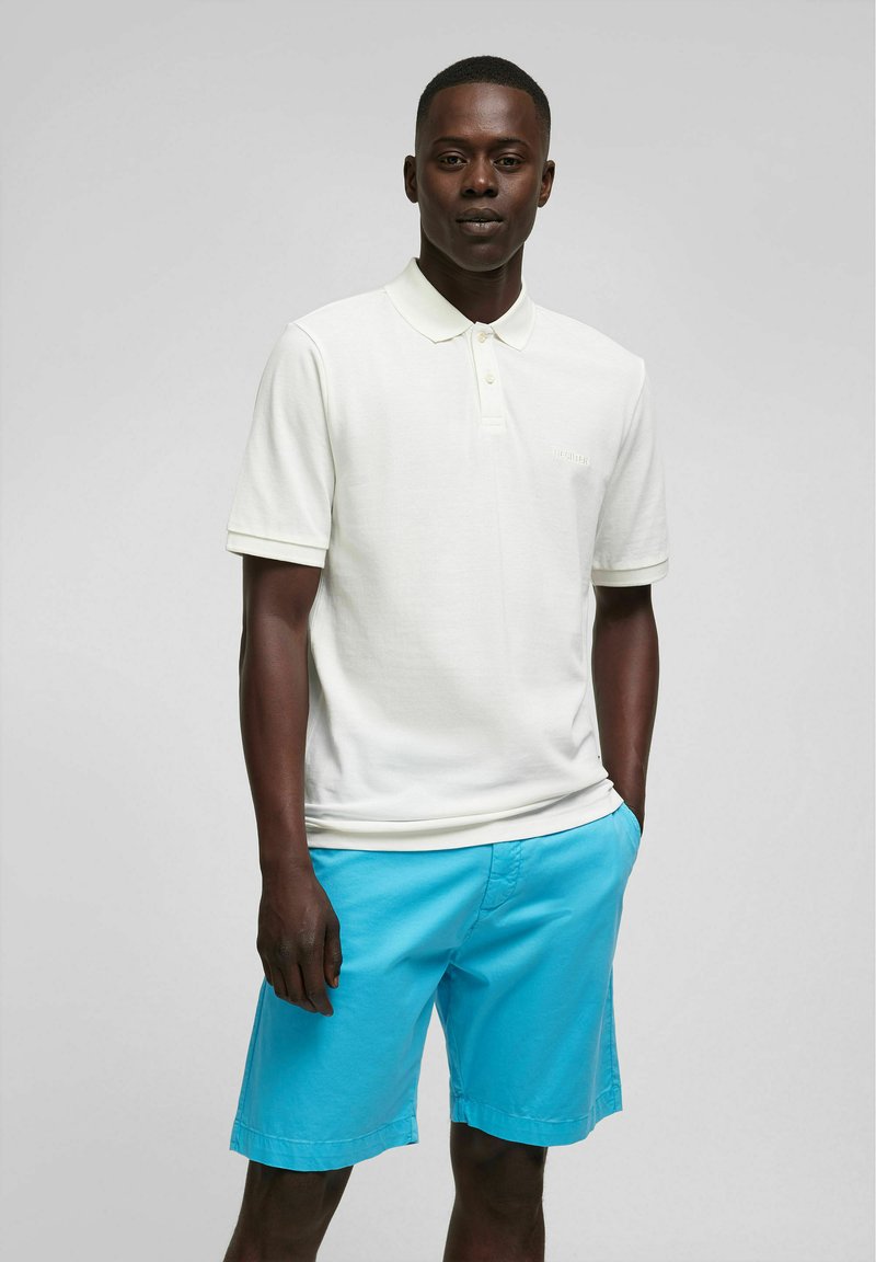 Witte poloshirt met een gestructureerde afwerking, korte mouwen en kraag, gecombineerd met lichtblauwe shorts, getoond op een model tegen een neutrale achtergrond.