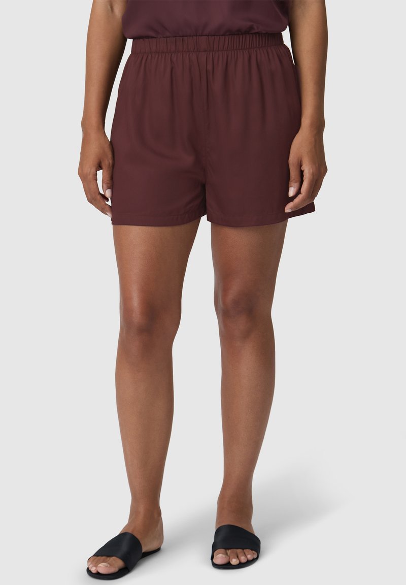 Pantalones cortos burdeos, ligeros, con una cintura elástica. Textura suave y diseño hasta la rodilla. Llevados con sandalias negras de dedo.