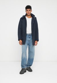 Parka imbottita blu marine con zip e chiusura a bottoni, dotata di cappuccio. Abbinata a jeans baggy azzurri e sneakers nere.