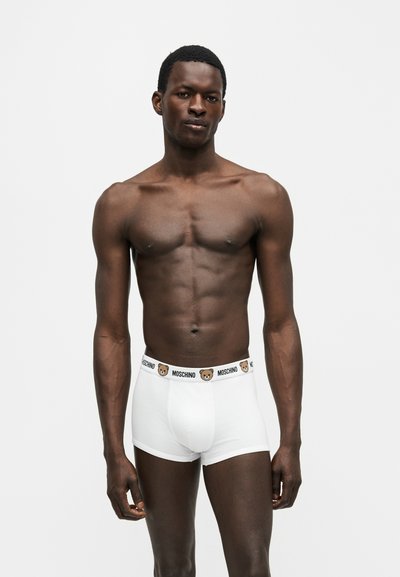 Homme torse nu portant un boxer blanc Moschino avec une ceinture à logo ours, debout devant un fond gris clair uni.
