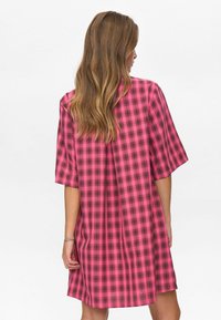 Vrouw met lang lichtbruin haar die een roze en zwart geruit kort mouwendraag jurk draagt, gezien van achteren op een witte achtergrond.