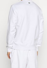 Sweat-shirt blanc en tissu doux, avec poignets et ourlet côtelés, arborant un petit logo vert à l'arrière du col. Design simple.