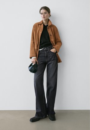 Junge Frau trägt eine braune Wildlederjacke, schwarzen Pullover, weite dunkle Jeans, hält eine kleine schwarze Handtasche und steht vor einer weißen Wand.
