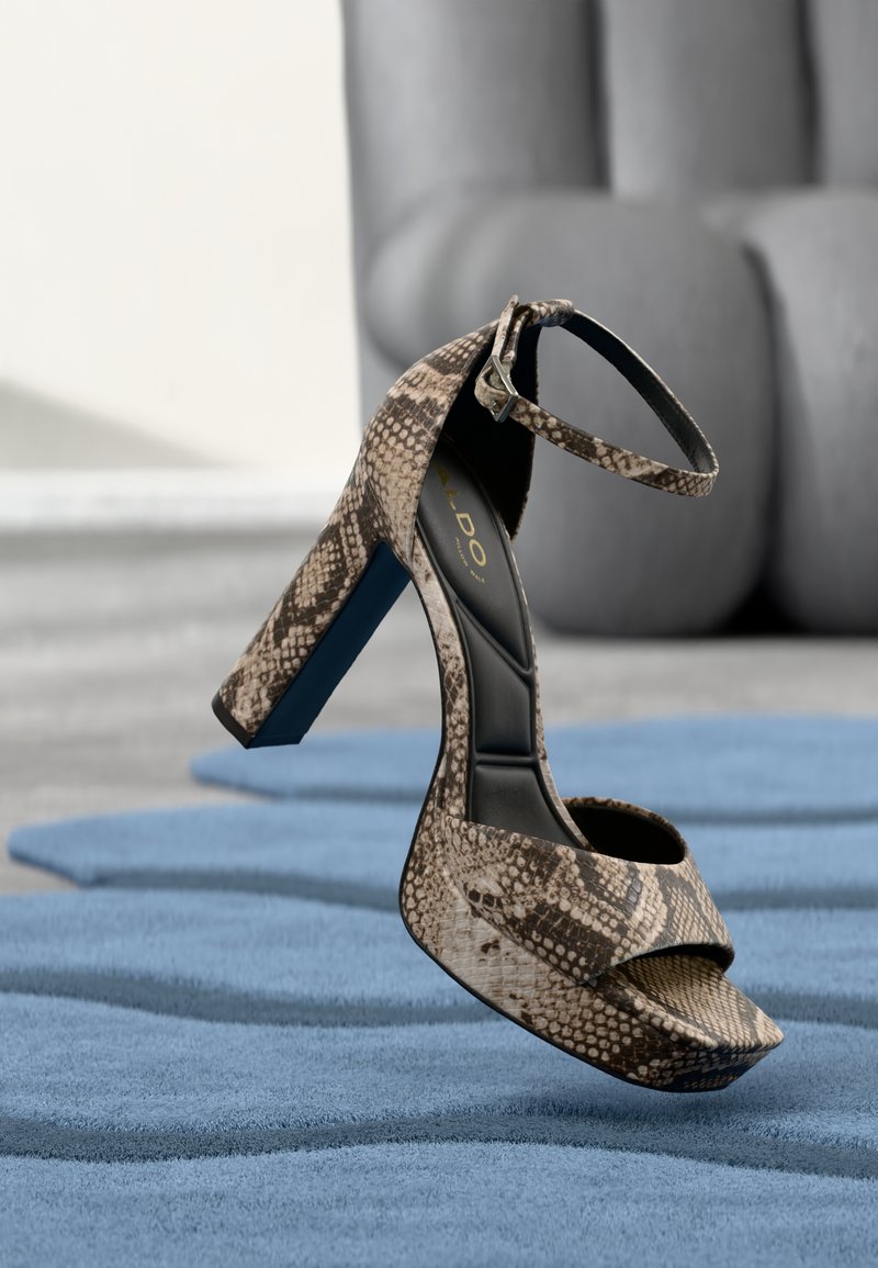 Kígyóbőr mintás magas sarkú szandál bokapánttal, kék texturált szőnyegen egyensúlyozva, elmosódott modern szürke szék háttérben.