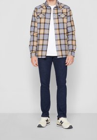 Camicia a quadri nei toni marrone, blu e beige, abbinata a jeans blu scuro e sneakers beige con logo nero.