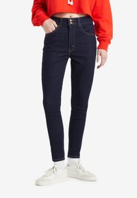 Sötét farmerból készült, magas derékú, két gombos záródással rendelkező skinny farmernadrág. Fehér sportcipőkkel és zoknikkal viselve.