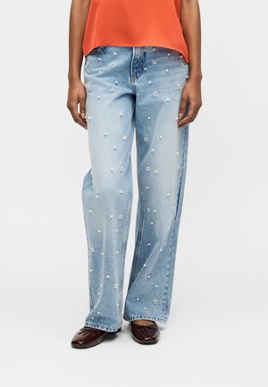 Jeans azzurri chiari con decorazioni di perle e fiori, indossati con scarpe rosso scuro e una maglia arancione, mostrati dalla vita ai piedi.